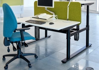 Adjustable operator task chair with inflatable lumbar support, arms and swivel base next to a back to back height adjustable desk with divider screen