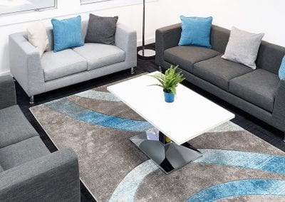 White top coffee table with chunky chrome leg and base, surrounded by grey low back sofas