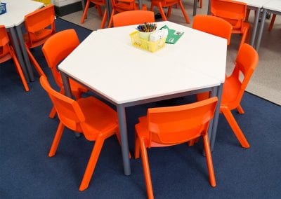 Hexagonal classroom table with white top and dark grey metal legs with orange plastic stacking chairs