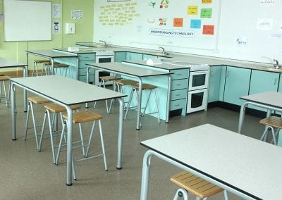 Educational high tables with white tops and grey metal legs, wood top stools and fully fitted kitchen training pods